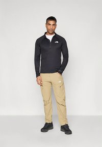 Juoda ilgomis rankovėmis užsegama palaidinė su užtrauktuku ir smėlio spalvos krovininės kelnės, derinamos su juodais žygių batais. Ant palaidinės ir kelnių yra „The North Face“ logotipai.