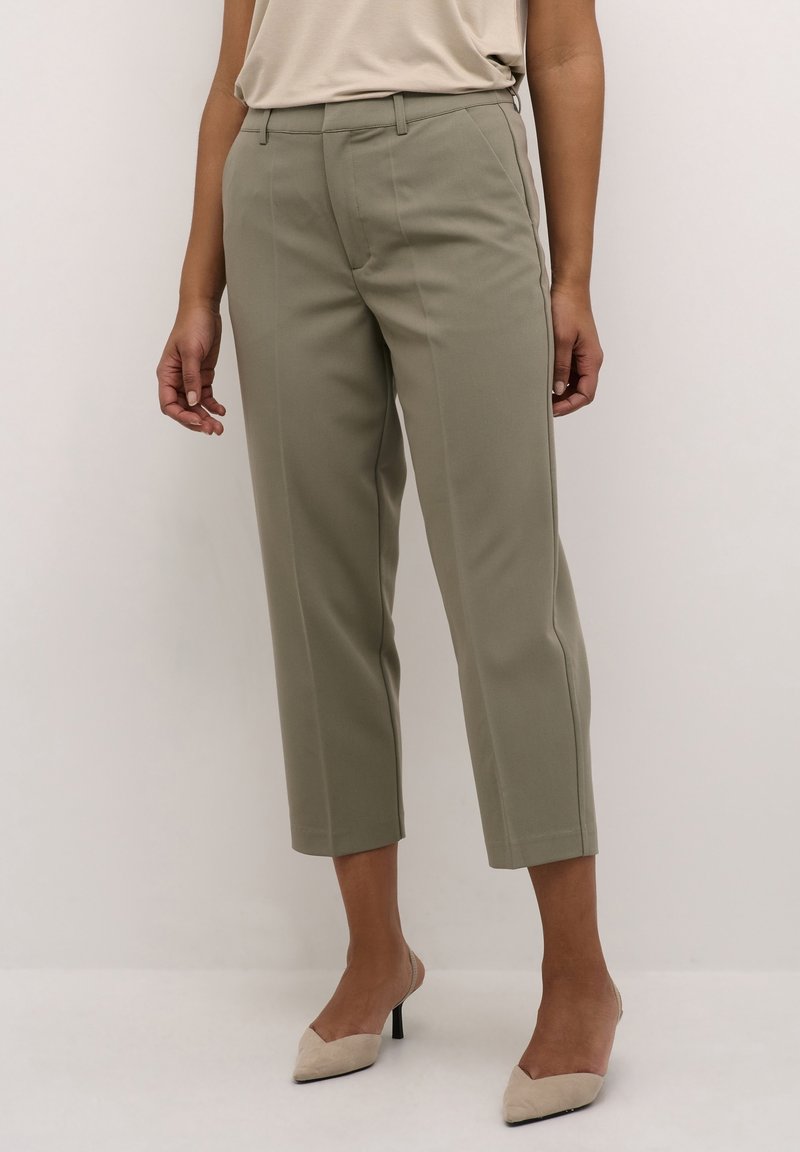 Femme portant un pantalon court taille haute vert olive avec des plis devant, associé à des chaussures à talons beige à bout pointu et bride arrière.