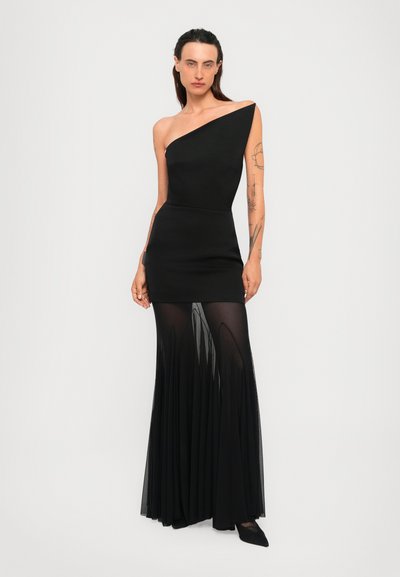 Kvinde iført en sort one-shoulder kjole med en tætsiddende mini nederdel og gennemskinnelig plisseret gulvlængde overlay, stående mod en ensfarvet baggrund.
