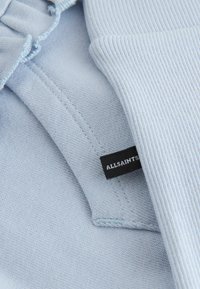 Material albastru deschis cu textură nervurată, având o etichetă inscripționată cu „ALLSAINTS” în text alb pe un fundal negru.