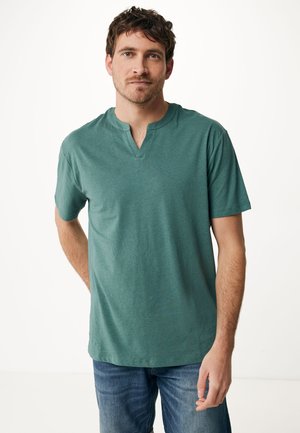 Camiseta verde de manga corta con un ligero escote en V, hecha de un tejido suave. Llevada con unos vaqueros azules, que presentan un ajuste casual y un detallado mínimo.