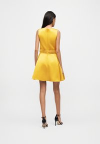 Γυναίκα που στέκεται με την πλάτη στραμμένη, φοράει ένα μαν sleeveless κίτρινο φόρεμα A-line με πτυχώσεις στη μέση και μαύρα ψηλοτάκουνα σανδάλια.