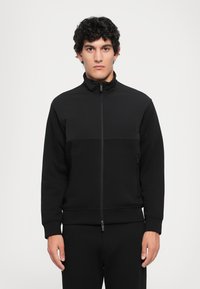Svart glidelåsjakke med høy krage, med en teksturert overdel og glatt nedre del. Stoffet har en matt finish.