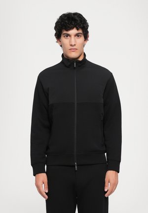 Veste noire zippée avec un col haut, présentant un panneau supérieur texturé et une section inférieure lisse. Le tissu a une finition mate.