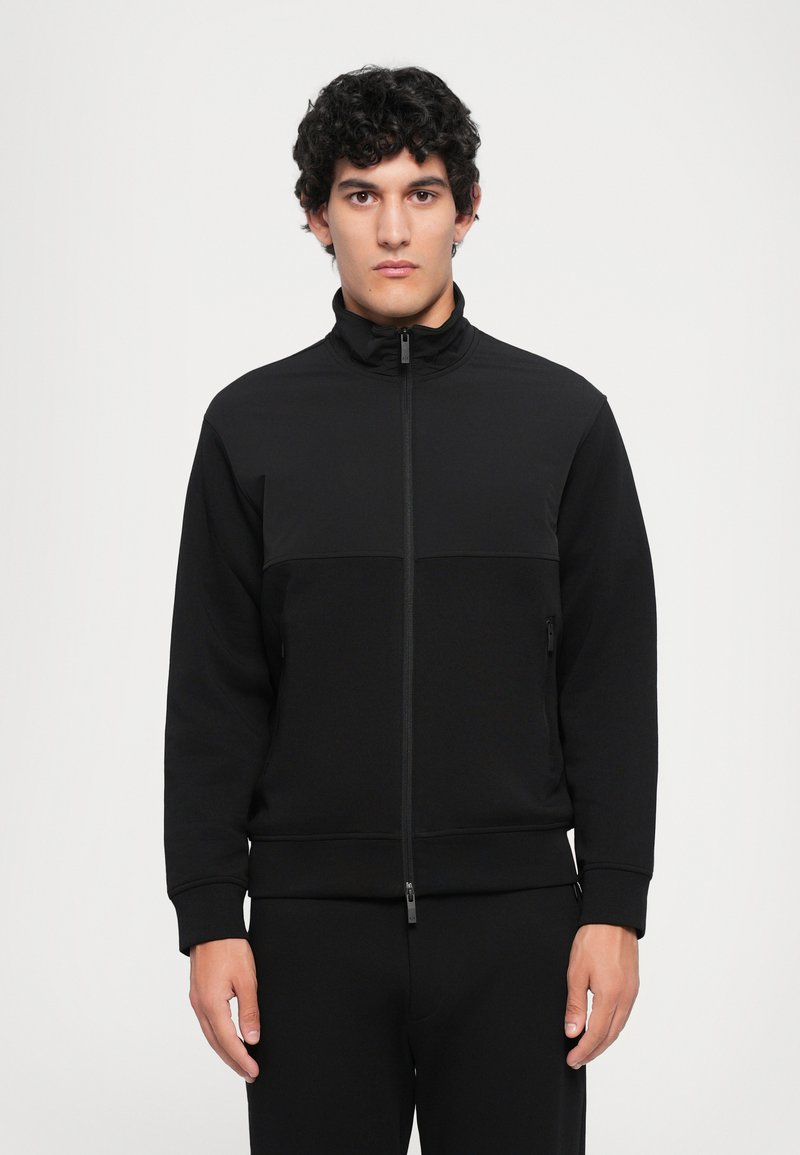 Svart glidelåsjakke med høy krage, med en teksturert overdel og glatt nedre del. Stoffet har en matt finish.