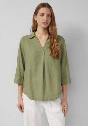 Kobieta o długich, jasnobrązowych włosach, ubrana w luźną oliwkowozieloną bluzkę z dekoltem w serek i rękawami do łokcia oraz białe spodnie na szarym tle.