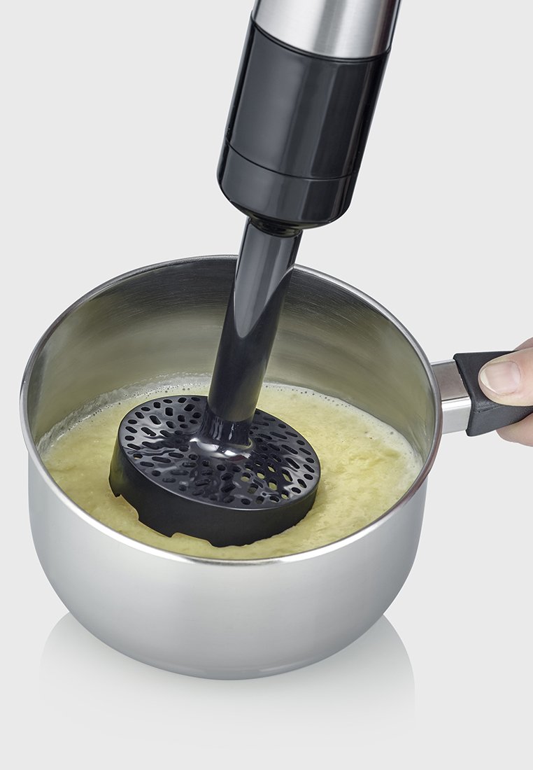 Eine Hand hält einen Edelstahltopf, während ein schwarzer Pürierstab mit Pürieraufsatz gelbes Essen im Topf püriert.