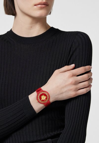 Red silicone watch with a circular gold dial featuring a lion motif and Greek key pattern. Worn on a woman's wrist, paired with a black ribbed top.