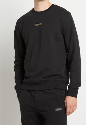 Homme portant un sweat-shirt à manches longues noir et un pantalon assorti, tous deux ornés de petits logos « HUGO » en lettres inversées sur la poitrine et la cuisse.