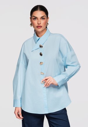 Femme portant une chemise bleu clair oversize avec de gros boutons argentés, un pantalon foncé et des boucles d'oreilles géométriques dorées, posant devant un fond uni.
