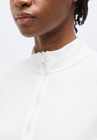 Κοντινό πλάνο ενός ατόμου που φορά λευκό ρούχο με φερμουάρ ψηλού γιακά, εμφανίζοντας τον λαιμό, το πηγούνι και μέρος του προσώπου και των μαλλιών.