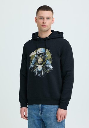 Jonge man draagt een zwarte hoodie met een afbeelding van een chimpansee die op een motor rijdt, gecombineerd met een blauwe spijkerbroek, staand tegen een effen achtergrond.