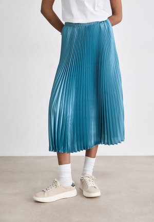 Personne portant une jupe midi plissée bleue, des chaussettes blanches, des baskets beiges et une chemise blanche, debout sur un sol clair.
