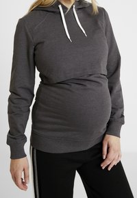 Szara bluza z kapturem o krótkim fasonie, wykonana z miękkiego materiału, z białymi sznurkami do ściągania i ściągaczami na rękawach, noszona na czarnych spodniach.