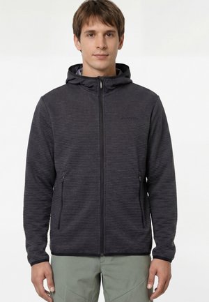 Jeune homme debout portant un sweat à capuche zippé gris foncé avec un motif à rayures horizontales subtil et un pantalon vert clair sur un fond uni.