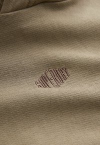 Kakifärgad tyg med en fin textur, med en broderad "Superdry"-logotyp i en mörkare nyans, belägen på en slät yta.