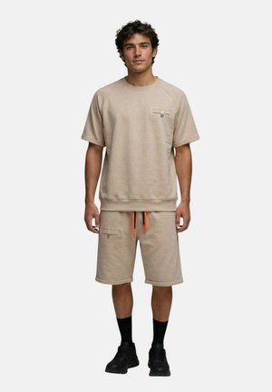 Homme debout face à l'avant portant un sweat-shirt beige à manches courtes et un short assorti avec des cordons orange, des chaussettes noires et des baskets noires.