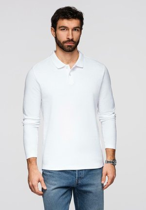 Mann mit dunklem Haar und Bart, der ein weißes Langarm-Poloshirt, blaue Jeans und eine Armbanduhr trägt, vor einem schlichten Hintergrund stehend.