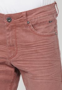 Gros plan d'un jean en denim rose délavé porté avec un haut blanc, montrant la poche avant, le bouton, les passants de ceinture et le tissu texturé.