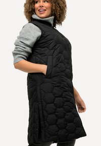 Schwarze gesteppte Weste mit hohem Kragen, seitlichen Taschen und Druckknopfverschlüssen, aus einem glatten, leichtgewichtigen Material gefertigt.