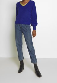 Blå ribbad stickad tröja med V-ringning, lager på lager över en gul topp med spetskant, tillsammans med högmidjade raka jeans och svarta stövlar.