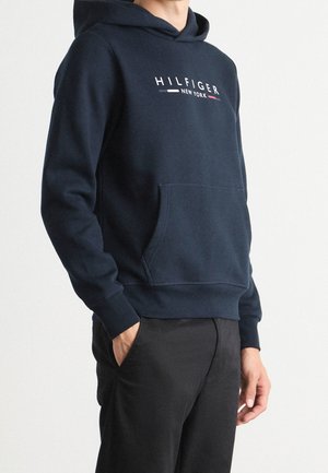 Osoba w granatowej bluzie z kapturem z napisem i logo "HILFIGER NEW YORK", czarne spodnie, stoi z jedną ręką w kieszeni.