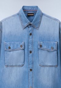 Ανοιχτό μπλε πουκάμισο από τζιν με μυτερό γιακά, εμπρόσθια σειρά κουμπιών και δύο τσέπες στο στήθος με καπάκι και μαύρα κουμπιά.