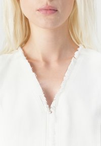 Witte kleding met een diepe V-halslijn met ruwe, rafelige randen, gemaakt van een textiel met structuur. Het ontwerp is eenvoudig en strak.
