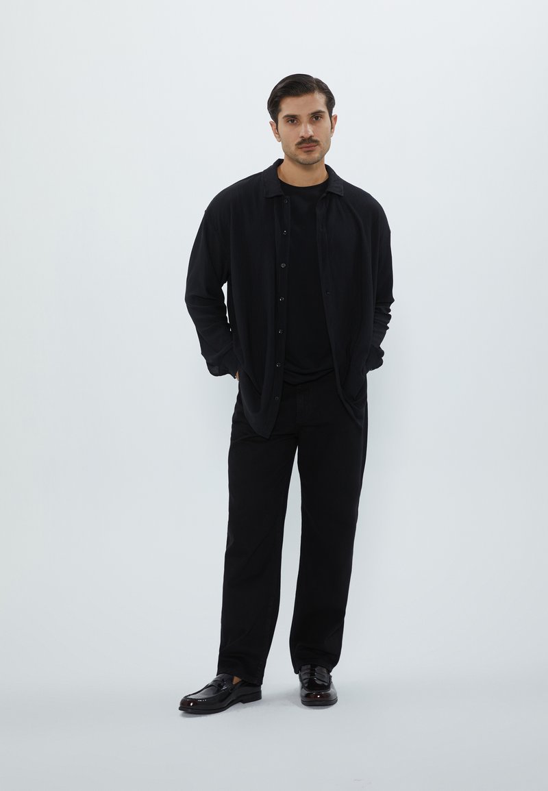 Chemise noire à boutons avec un motif texturé, associée à un t-shirt noir et un pantalon noir ; des chaussures noires brillantes complètent le look.