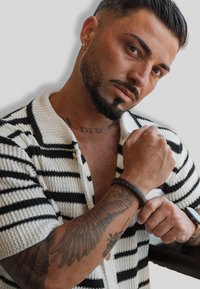Knit short-sleeve cardigan in white and black horizontal stripes, featuring a collar and textured fabric. Bracelet and tattoo visible on arm.