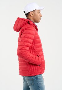 Rød polstret jakke med hætte, der har vandrette quiltede sektioner og sidelommer. Bæres over en blå ternet skjorte og jeans.