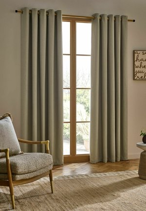 Beige Vorhänge hängen an einer Holzstange über einem hohen Fenster mit Gartenblick, neben einem gepolsterten Stuhl und einem runden Tisch mit einer Pflanze.