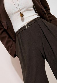 Bruine geplooide broek van een gestructureerde stof met zilverkleurige accenten bij de taille. Gecombineerd met een wit shirt en een donker jasje.