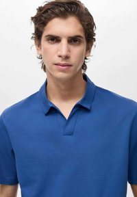 Sininen polo-paita, jossa on klassinen kaula-aukko, lyhyet hihat, teksturoitu kangas ja yksi napitus kaula-aukolla.