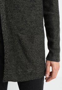 Mörkgrå stickad cardigan med en texturerad tyg, öppen fram och en framficka. Ärmsluten är ribbade för en figurnära look.