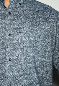Camicia a maniche lunghe con fantasia floreale in azzurro chiaro e disegni blu scuro. Presenta un colletto e bottoni neri. Piccolo logo sulla tasca del petto.