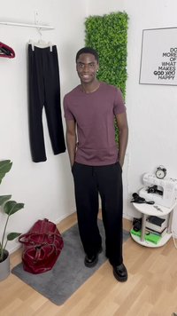 Bordeauxfarbendes Kurzarmhemd, schwarze weite Hose und klobige schwarze Schuhe. Rote Tasche und Pflanzen in einem stilvollen Raum mit einer Nähmaschine.