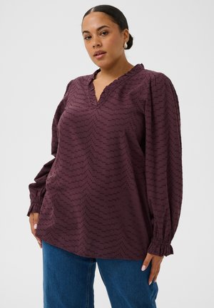Femme aux cheveux foncés en chignon bas, portant des boucles d'oreilles anneaux dorés, un chemisier violet texturé à manches longues et un jean bleu, debout devant un fond uni.