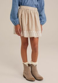 Világos bézs szoknya hímzett virágmintás díszítéssel, elasztikus derékpánttal és réteges kivitelezéssel, szürke bokacsizmával és fehér fodros zoknikkal párosítva.