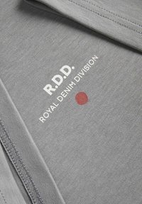 Pelēks kokvilnas audums ar baltu tekstu "R.D.D. ROYAL DENIM DIVISION" un mazu sarkanu apli. Gluda tekstūra ar redzamām šuvēm.
