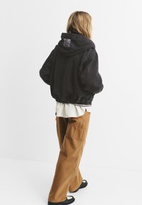Jachetă neagră căptușită cu glugă, cu hem și mansete elasticizate; suprapusă peste o cămașă albă texturată și pantaloni maro cu croi larg. Sandale negre.