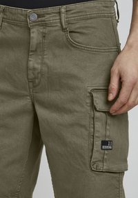 Olijfgroene cargobroek met een losse pasvorm, voorzien van een zijzak, gestikte details en een zwarte logopatch op de linker dij.