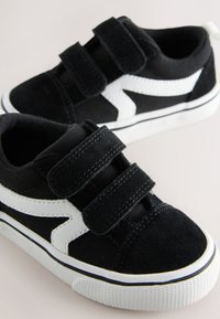Baskets noires pour enfants avec des bandes blanches en zigzag sur le côté et deux bandes velcro noires sur fond clair.