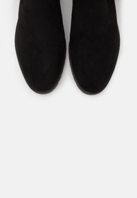 Botines de ante negros con una textura suave, puntera redondeada y suela plana de goma, exhibiendo un diseño minimalista y líneas limpias.