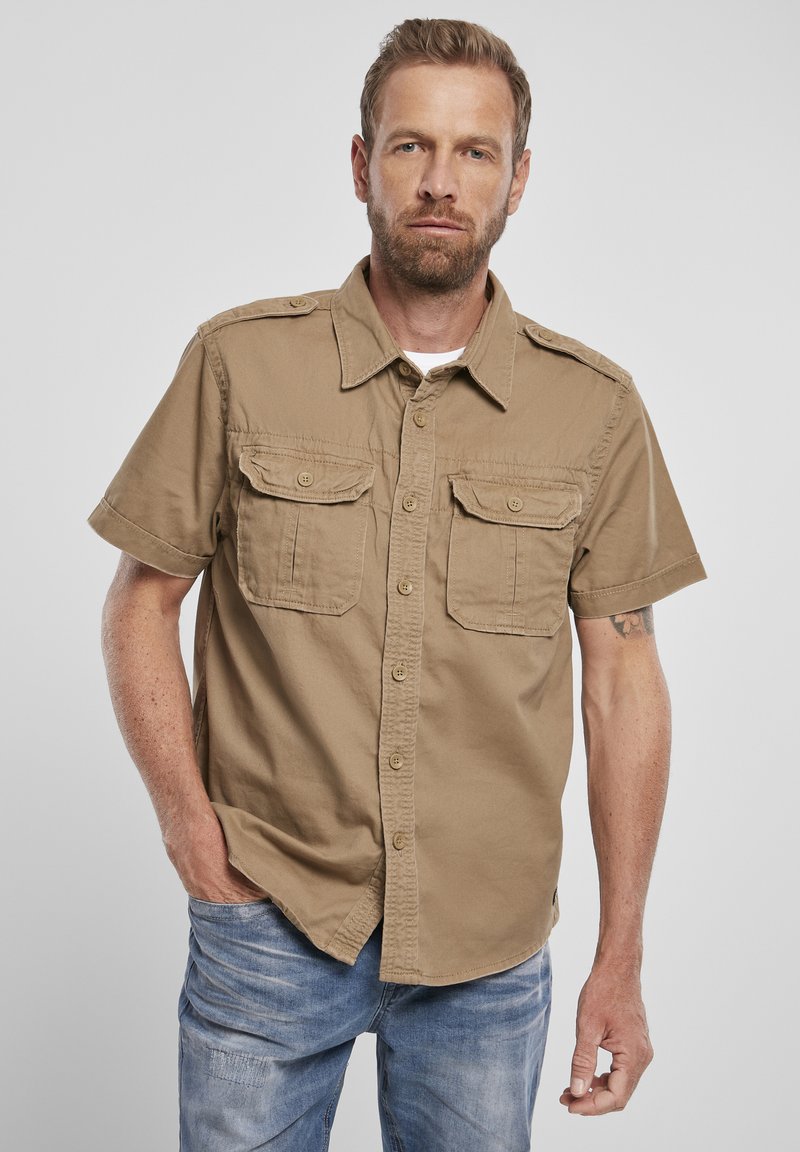 Camicetta button-up khaki a maniche corte con due tasche sul petto, colletto e una leggera texture; indossata con jeans azzurri chiari.