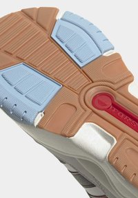 Sula till sko med texturerade greppsektioner i ljusblått och brunt, en röd detalj med präglad logotyp och vit mellansula synlig.