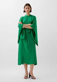 Vihreä silkki mekko, jossa on korkea pääntie ja kimono-tyyliset hihat, näyttäen löysältä ja sileältä, yhdistettynä mustiin hihna-korkokenkiin.