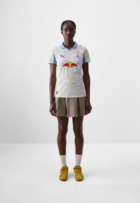 T-shirt à manches courtes de couleur bleu pâle avec des motifs floraux, arborant un logo rouge et des accents bleus ; associé à une jupe plissée marron et des chaussures jaunes.