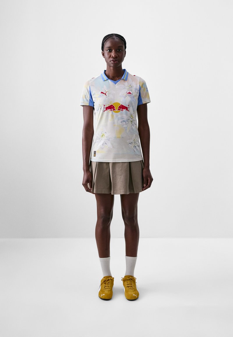 T-shirt à manches courtes de couleur bleu pâle avec des motifs floraux, arborant un logo rouge et des accents bleus ; associé à une jupe plissée marron et des chaussures jaunes.