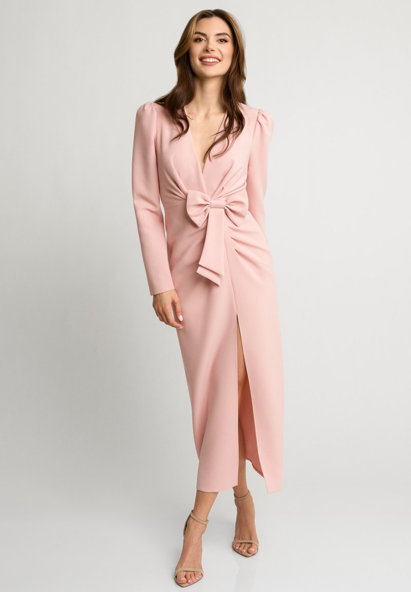 Różowa sukienka z długim rękawem, z głębokim dekoltem w kształcie litery V, z detalem w postaci kokardy z przodu, marszczeniami oraz wysokim rozcięciem, wykonana z gładkiego materiału.